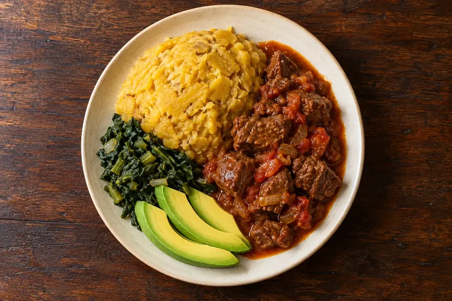 Overhead view of a plate with mashed matooke, a tomato stew, cooked greens and sliced avocado on a wooden table